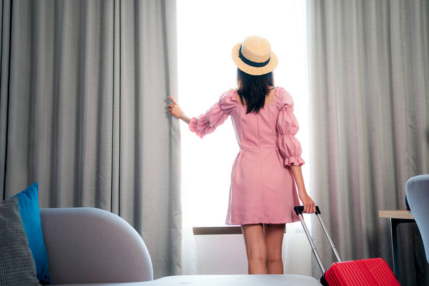 Путешественница азиатского происхождения в розовом платье прибывает в номер в отеле и открывает занавеску, чтобы насладиться видом снаружи, это изображение может быть использовано для путешествия, тура, отеля и страхования путешествий концепции - Фото, изображение