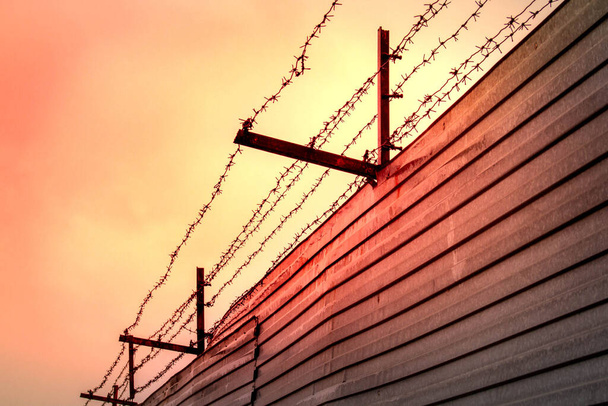 Alambre de púas estirado sobre soportes oxidados montados en una cerca hecha de láminas de metal contra el telón de fondo del cielo rojo atardecer - Foto, Imagen