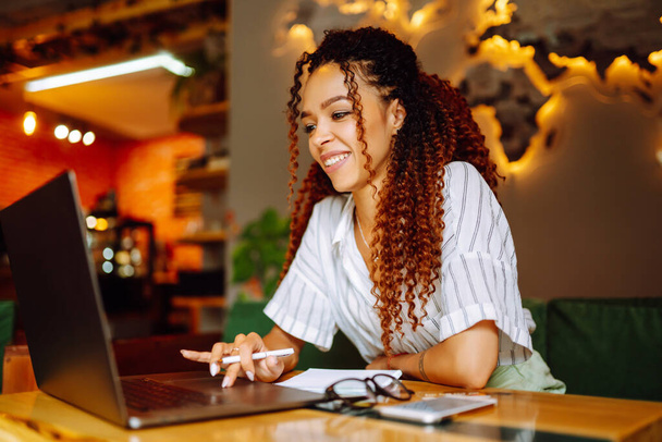 Young freelancer woman using laptop computer sitting at cafe table. Smiling woman working online or studying and learning while using notebook. Business People Concept - 写真・画像