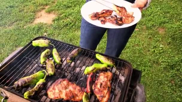 A woman is standing by a small charcoal grill where she grills marinated chicken and shishito peppers. She flips them with tongs and takes the ones that are ready onto a white plate. - Footage, Video