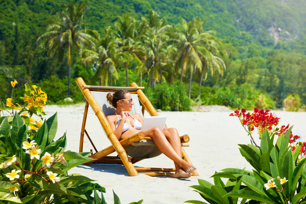 Mujer feliz con portátil en silla de bambú trabajando remotamente en la playa tropical - Foto, Imagen