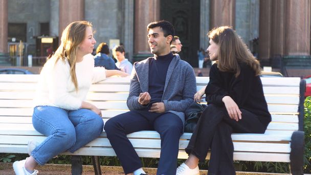 Dos chicas jóvenes y un hombre amigos sentados en un banco en el parque de verano charlando felizmente divertirse. Medios. Retrato de dos mujeres sonrientes y un hombre pasando tiempo juntos y hablando. - Foto, imagen