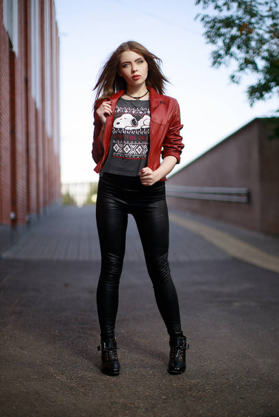 Portret van een jonge vrouw in een rood leren jasje en zwarte legging blijven overdag in de stad. - Foto, afbeelding