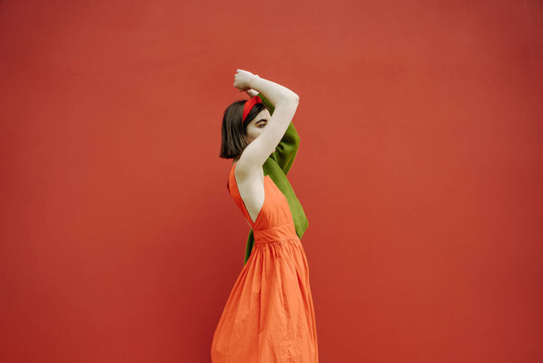 a slender touching thin girl stands sideways in a red dress and a green jacket. She raised her hands dreamily. unrecognizable portrait no face. Romantic mood. red wall background. hope in future - Foto, Bild