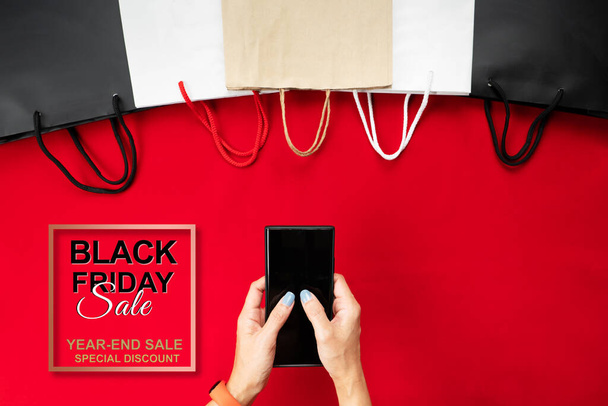 ブラック・フライデーセール赤い背景にショッピングバッグ付きスマートフォンで女性の手オンラインショッピング - 写真・画像