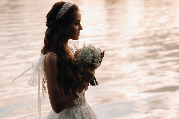 An elegant bride in a white dress and gloves stands by the river in the Park with a bouquet, enjoying nature at sunset.A model in a wedding dress and gloves in a nature Park.Belarus. - Φωτογραφία, εικόνα