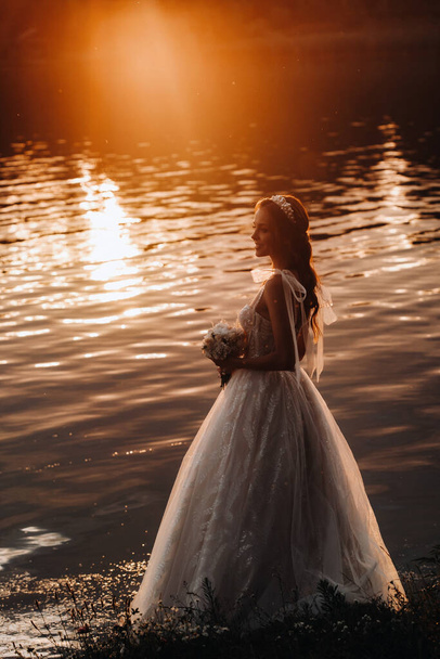 Μια κομψή νύφη σε ένα λευκό φόρεμα και γάντια στέκεται δίπλα στο ποτάμι στο πάρκο με ένα μπουκέτο, απολαμβάνοντας τη φύση στο ηλιοβασίλεμα. Ένα μοντέλο σε ένα νυφικό και γάντια σε ένα πάρκο φύσης.Λευκορωσία. - Φωτογραφία, εικόνα