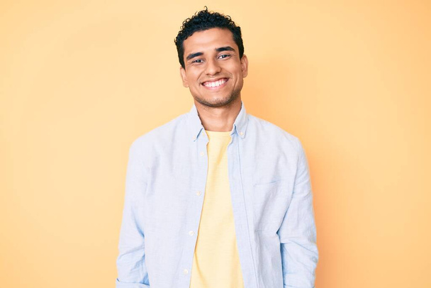 Joven hombre hispano guapo de pie sobre fondo amarillo con una sonrisa alegre y fresca en la cara. persona afortunada.  - Foto, Imagen