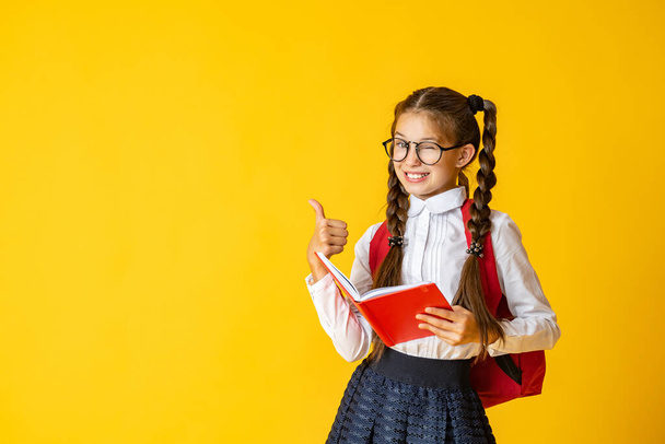 cute 10-year-old Caucasian schoolgirl with glasses and a backpack gives a thumbs up in approval and winks her eye. Returns to school. The child smiles and poses on a yellow background. Back to school - Фото, изображение