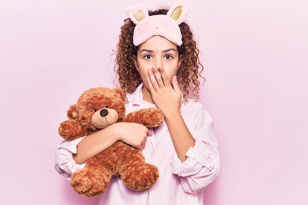 Beautiful kid girl with curly hair wearing sleep mask and pajamas holding teddy bear covering mouth with hand, shocked and afraid for mistake. surprised expression  - Foto, Imagem