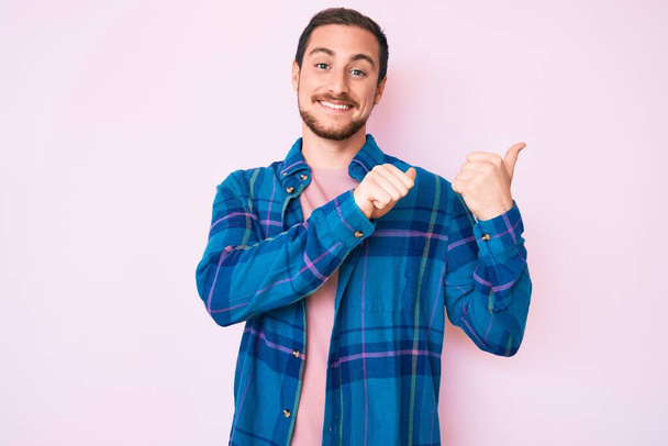 Mladý pohledný muž v neformálních šatech ukazuje na záda s rukou a palci nahoru, sebevědomě se usmívá  - Fotografie, Obrázek