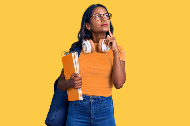 Joven chica india sosteniendo mochila de estudiante y libros cara seria pensando en la pregunta con la mano en la barbilla, reflexivo acerca de la idea confusa  - Foto, Imagen