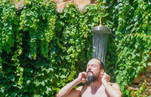 Parrakas hipsteri mies ottaa ulkona suihku puutarhassa kasvien kanssa - Valokuva, kuva