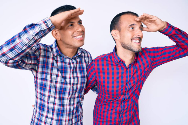 Joven pareja gay de dos hombres vistiendo ropa casual muy feliz y sonriente mirando lejos con la mano sobre la cabeza. concepto de búsqueda.  - Foto, imagen