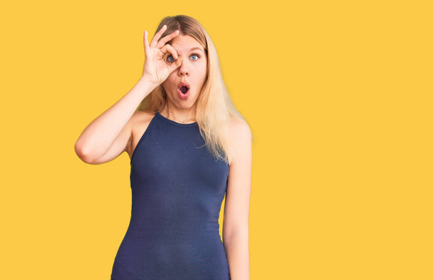 Young beautiful blonde woman wearing casual dress doing ok gesture shocked with surprised face, eye looking through fingers. unbelieving expression.  - Photo, Image