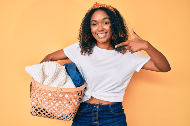 Joven mujer afroamericana sosteniendo cesta de la ropa sonriendo feliz señalando con la mano y el dedo  - Foto, Imagen