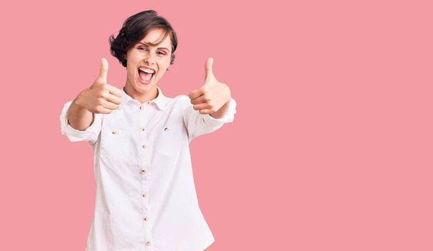 Beautiful young woman with short hair wearing elegant white shirt approving doing positive gesture with hand, thumbs up smiling and happy for success. winner gesture.  - Фото, изображение