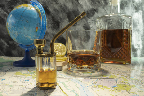 Großaufnahme einer Bong mit Marihuana und einer Flasche Whiskey auf einem Tisch mit einer Weltkugel und einer Lupe - Foto, Bild