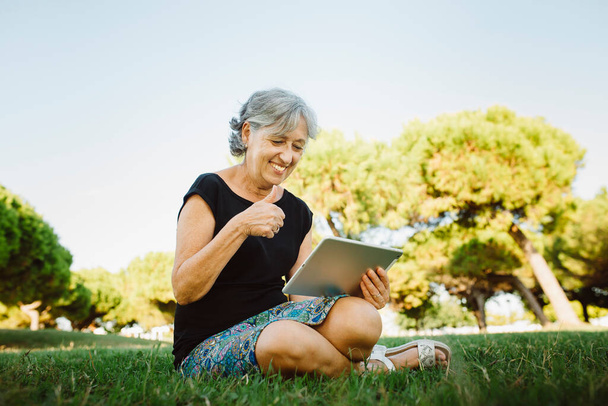 Mujer mayor charlando en línea con su tableta en un parque - Foto, Imagen