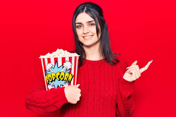 Young beautiful girl eating popcorn smiling happy pointing with hand and finger to the side  - Fotoğraf, Görsel