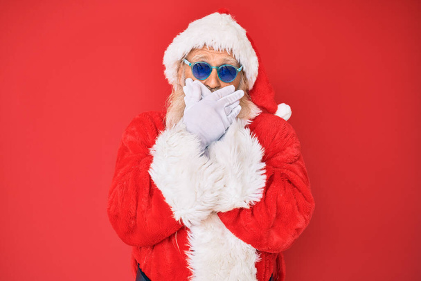 Ein alter älterer Mann in Weihnachtsmann-Kostüm und Sonnenbrille schockierte, als er fälschlicherweise den Mund mit den Händen verdeckte. Geheimes Konzept.  - Foto, Bild