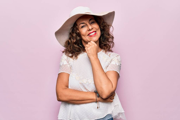 Μέση ηλικία όμορφη γυναίκα φορώντας καλοκαιρινό t-shirt και καπέλο πάνω από απομονωμένο ροζ φόντο χαμογελώντας αναζητούν αυτοπεποίθηση στην κάμερα με σταυρωμένα χέρια και το χέρι στο πηγούνι. Θετική σκέψη. - Φωτογραφία, εικόνα