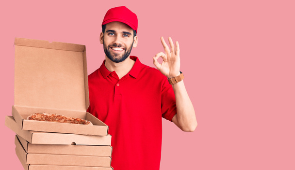 Joven hombre guapo con barba llevando uniforme de entrega sosteniendo cajas con pizza haciendo signo de ok con los dedos, sonriendo gesto amistoso excelente símbolo  - Foto, imagen