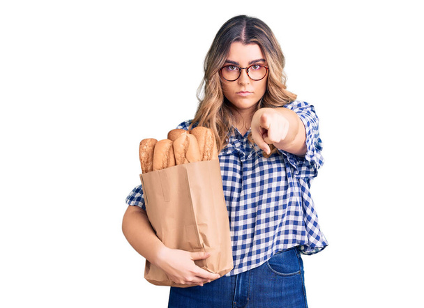Joven mujer caucásica sosteniendo bolsa de papel con pan señalando con el dedo a la cámara y a ti, gesto confiado mirando en serio  - Foto, imagen