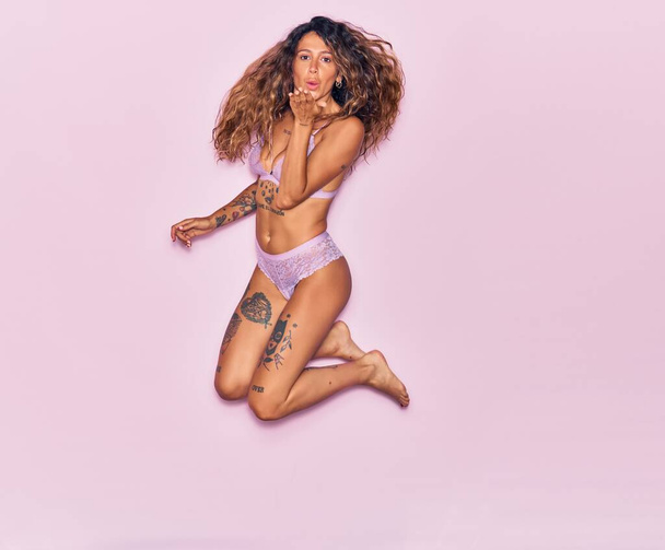Young beautiful curly woman with tattoo wearing lingerie smiling happy. Jumping with smile on face throwing kiss with hands over isolated pink background - Foto, Bild