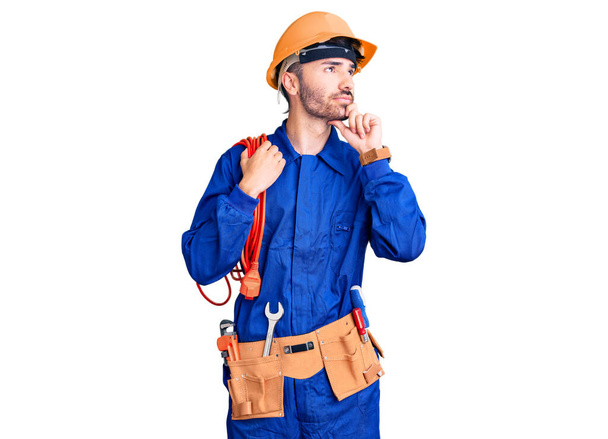 Jovem hispânico vestindo uniforme eletricista segurando cabo rosto sério pensando em questão com a mão no queixo, pensativo sobre ideia confusa  - Foto, Imagem