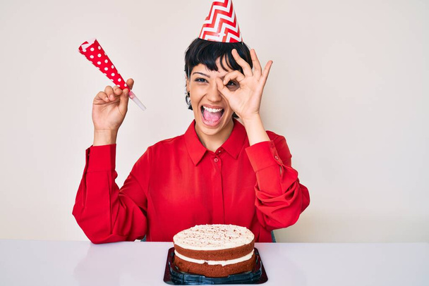 Красива брюнетка жінка з коротким волоссям, що святкує день народження з тортами, посміхаючись, щаслива, роблячи знак ОК з рукою на око дивиться крізь пальці
  - Фото, зображення