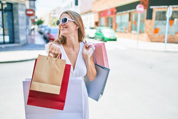 Fiatal szőke nő mosolyog boldog gazdaság bevásárló táskák az utcán a város - Fotó, kép