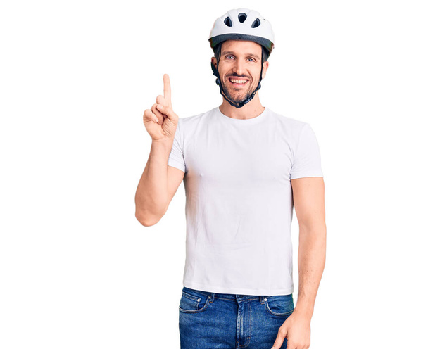 Молодий красивий чоловік у велосипедному шоломі показує і вказує пальцем номер один, посміхаючись впевнено і щасливо
.  - Фото, зображення