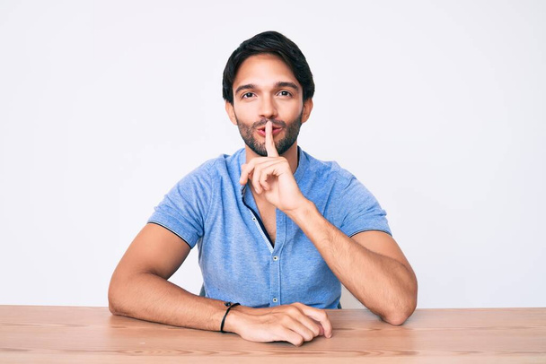 Handsome hispanic man wearing casual clothes sitting on the table asking to be quiet with finger on lips. silence and secret concept.  - Photo, Image