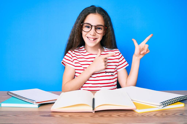 Menina hispânica bonito estudando para o exame escolar sentado na mesa sorrindo e olhando para a câmera apontando com duas mãos e dedos para o lado.  - Foto, Imagem