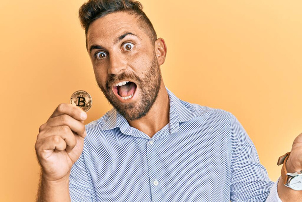 Handsome man with beard holding virtual currency bitcoin celebrating victory with happy smile and winner expression with raised hands  - Fotoğraf, Görsel