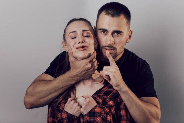 The man holds the woman by the neck and ordered to be silent. Scared woman with bruises on her face. Domestic violence, victim of violence - Фото, изображение