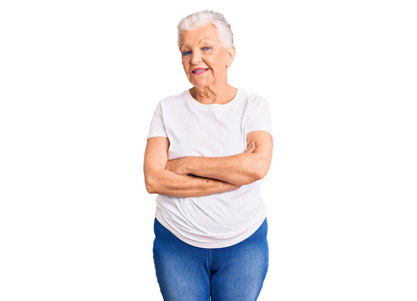 Eine ältere, schöne Frau mit blauen Augen und grauen Haaren trägt ein lässiges weißes T-Shirt und lächelt mit verschränkten Armen in die Kamera. positive Person.  - Foto, Bild