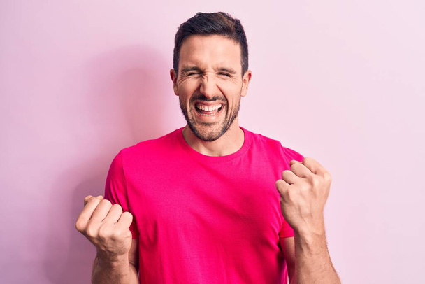 Joven hombre guapo con camiseta casual de pie sobre un fondo rosa aislado celebrando sorprendido y sorprendido por el éxito con los brazos levantados y los ojos cerrados - Foto, imagen