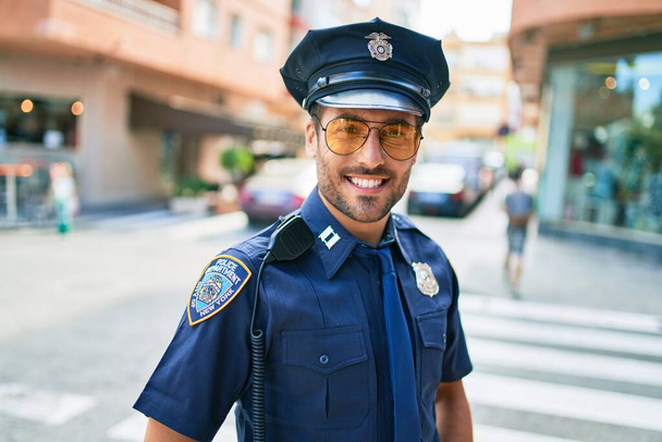Junger gut aussehender hispanischer Polizist in Polizeiuniform, der glücklich lächelnd an der Stadtstraße steht. - Foto, Bild