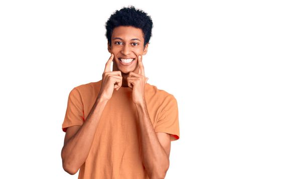 Молодой африканский американец в повседневной одежде, улыбающийся с открытым ртом, указывающий пальцами и заставляющий веселую улыбку  - Фото, изображение