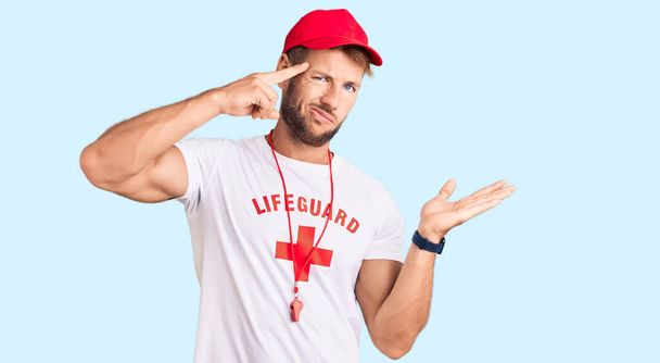 Young caucasian man wearing lifeguard t shirt holding whistle confused and annoyed with open palm showing copy space and pointing finger to forehead. think about it.  - Photo, Image