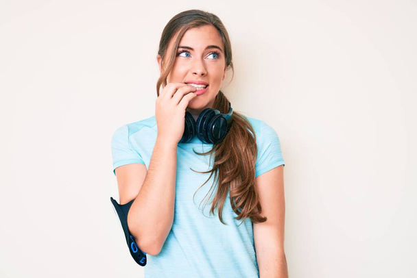 Beautiful young caucasian woman wearing gym clothes and using headphones looking stressed and nervous with hands on mouth biting nails. anxiety problem.  - Photo, Image