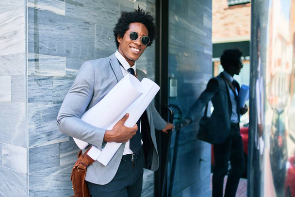 Fiatal jóképű afro-amerikai építész, aki boldogan mosolyog. Mosollyal az arcán, tervrajzokat tartva az iroda ajtajában.. - Fotó, kép