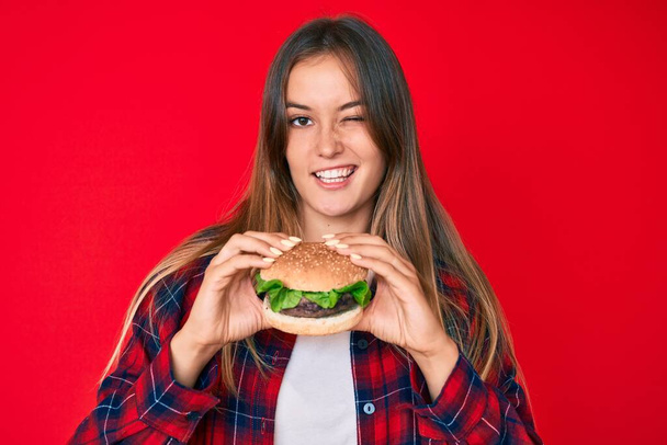 Gyönyörű kaukázusi nő eszik egy ízletes klasszikus burger kacsintás néz a kamera szexi kifejezés, vidám és boldog arc.  - Fotó, kép
