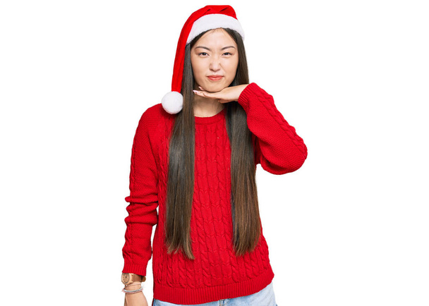 Young chinese woman wearing christmas hat cutting throat with hand as knife, threaten aggression with furious violence  - Photo, Image