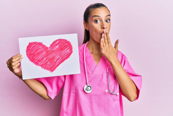Jeune femme hispanique portant l'uniforme de médecin tenant le coeur dessiner couvrant la bouche avec la main, choqué et peur de l'erreur. expression surprise  - Photo, image
