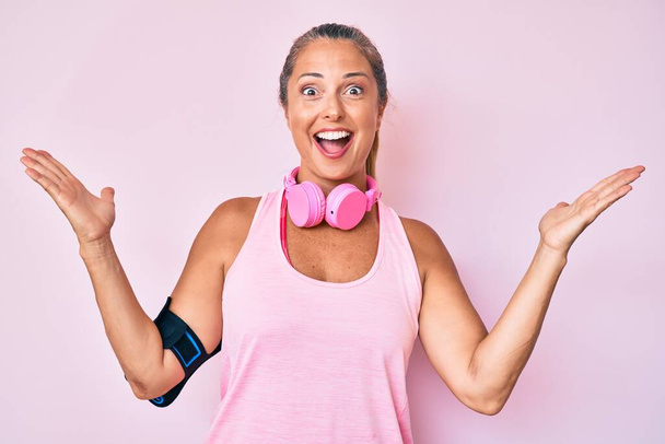 Латиноамериканка средних лет в спортивной одежде и с помощью наушников празднует победу с счастливой улыбкой и выражением победителя с поднятыми руками  - Фото, изображение