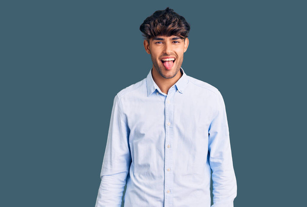 Joven hombre hispano con camisa casual sacando la lengua feliz con expresión divertida. concepto de emoción.  - Foto, Imagen