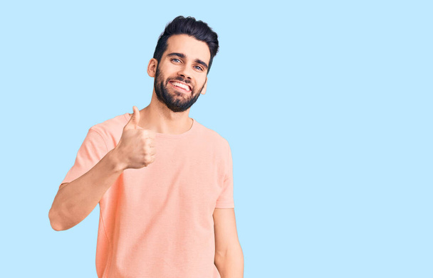 Mladý pohledný muž s plnovousem, který nosí neformální tričko a dělá šťastné gesto rukou nahoru. schválení výrazu při pohledu na fotoaparát, který vykazuje úspěch.  - Fotografie, Obrázek